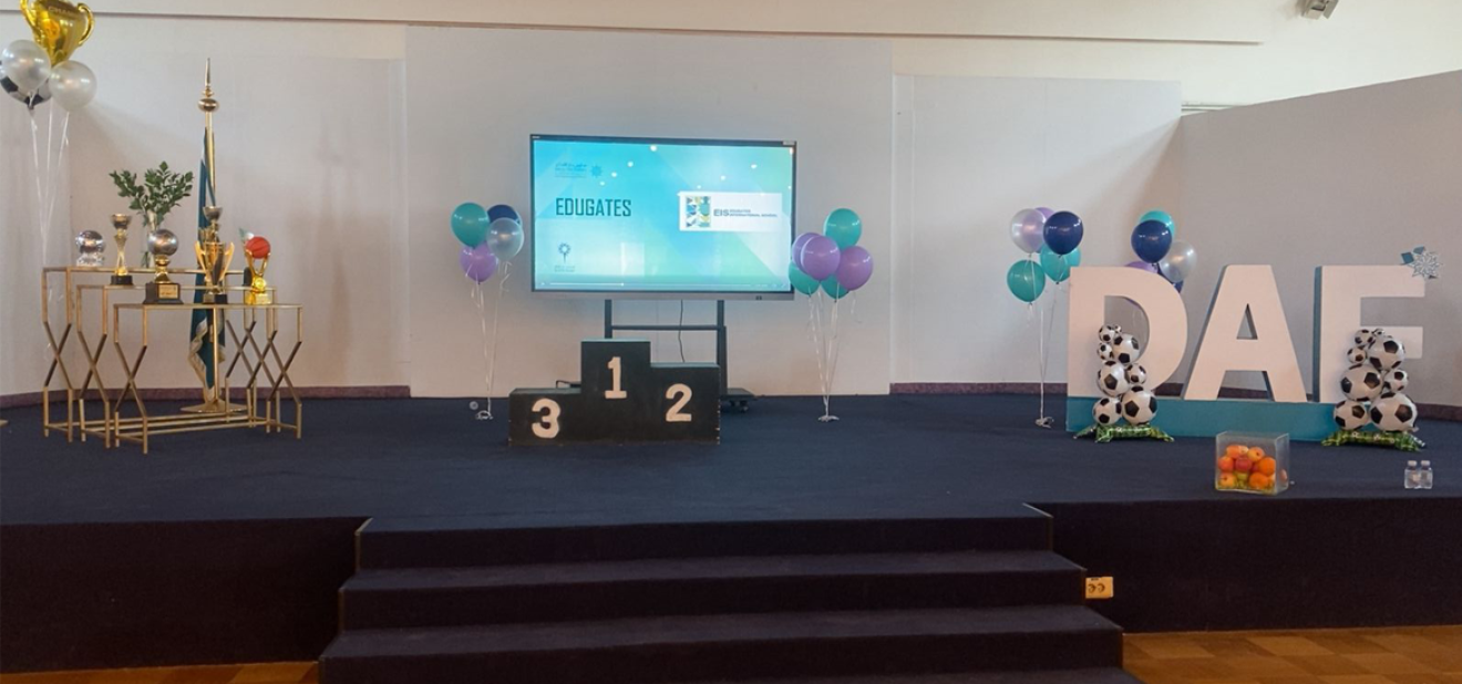 منصة مزينة لفعالية أولمبياد دار الفكر الرياضي 2025، تحتوي على جوائز وكؤوس، منصة تتويج بالمراكز 1 و2 و3، بالونات بألوان المدرسة، شاشة عرض، وحروف DAF كبيرة مزينة بكرات القدم.